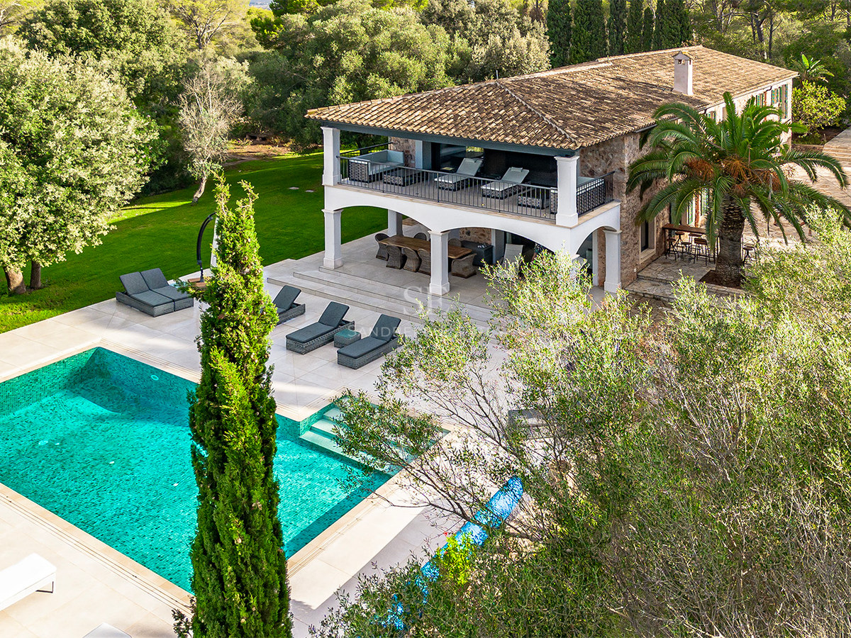 Vista elevada de una villa de piedra con piscina turquesa, tumbonas y jardines mediterráneos frondosos.