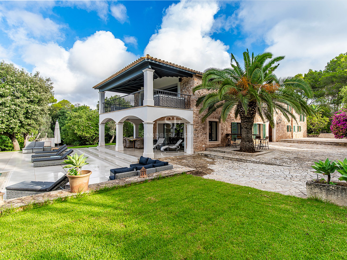 Villa de piedra natural de dos niveles con arcos, palmera central, jardín cuidado y zona de piscina.