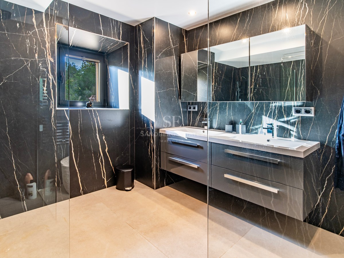 Baño moderno con paredes de mármol negro veteado, mueble de doble lavabo gris y ducha con mampara de cristal.