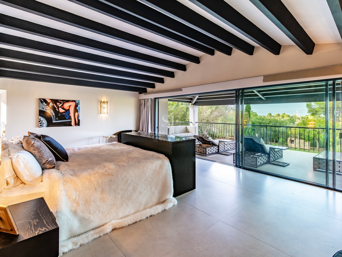 Dormitorio principal moderno con vigas oscuras y puertas de cristal que dan a una terraza con vistas al bosque.