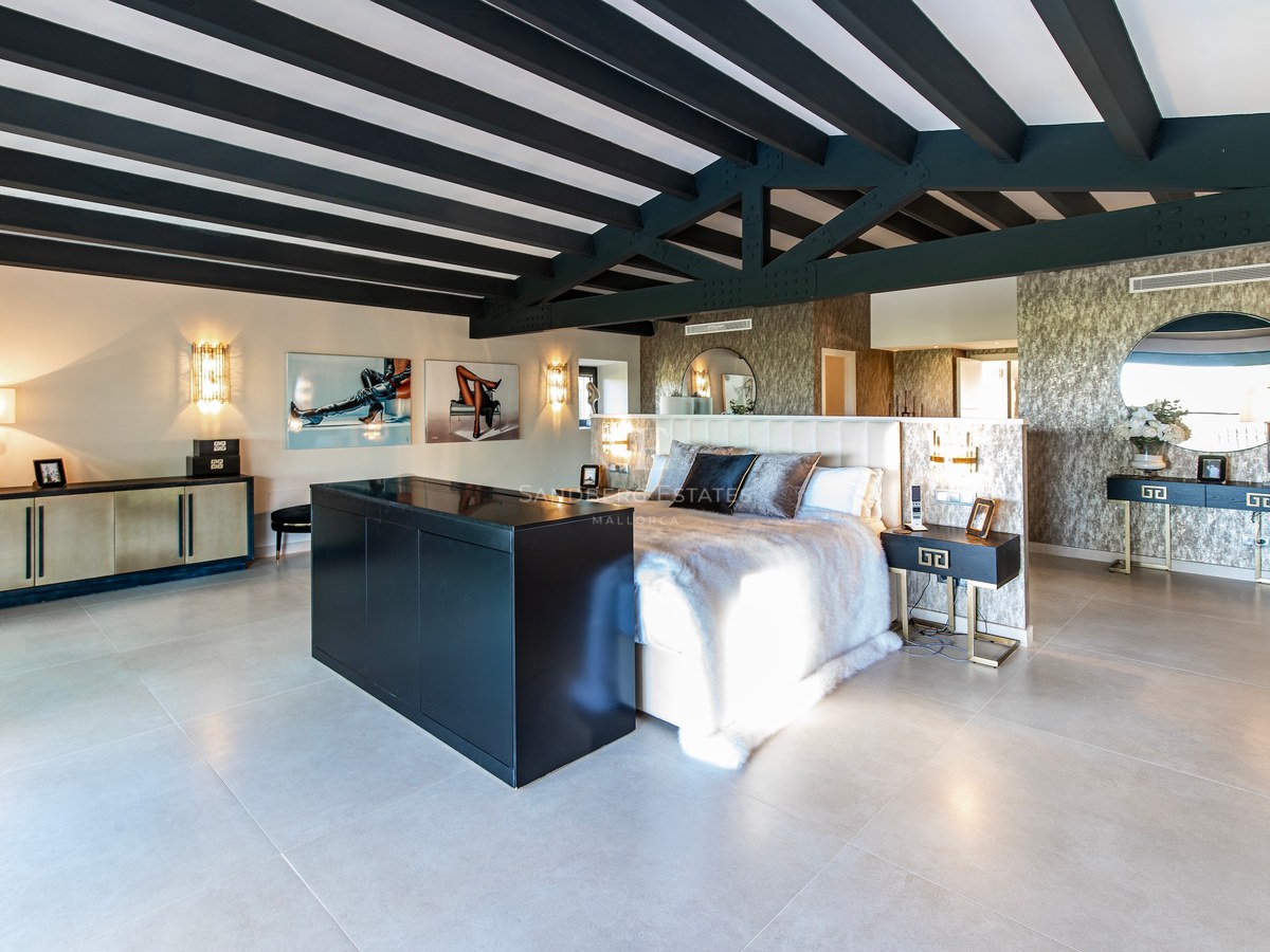 Amplio dormitorio principal con vigas de madera negras, cama king con manta de piel y suelos de piedra clara.