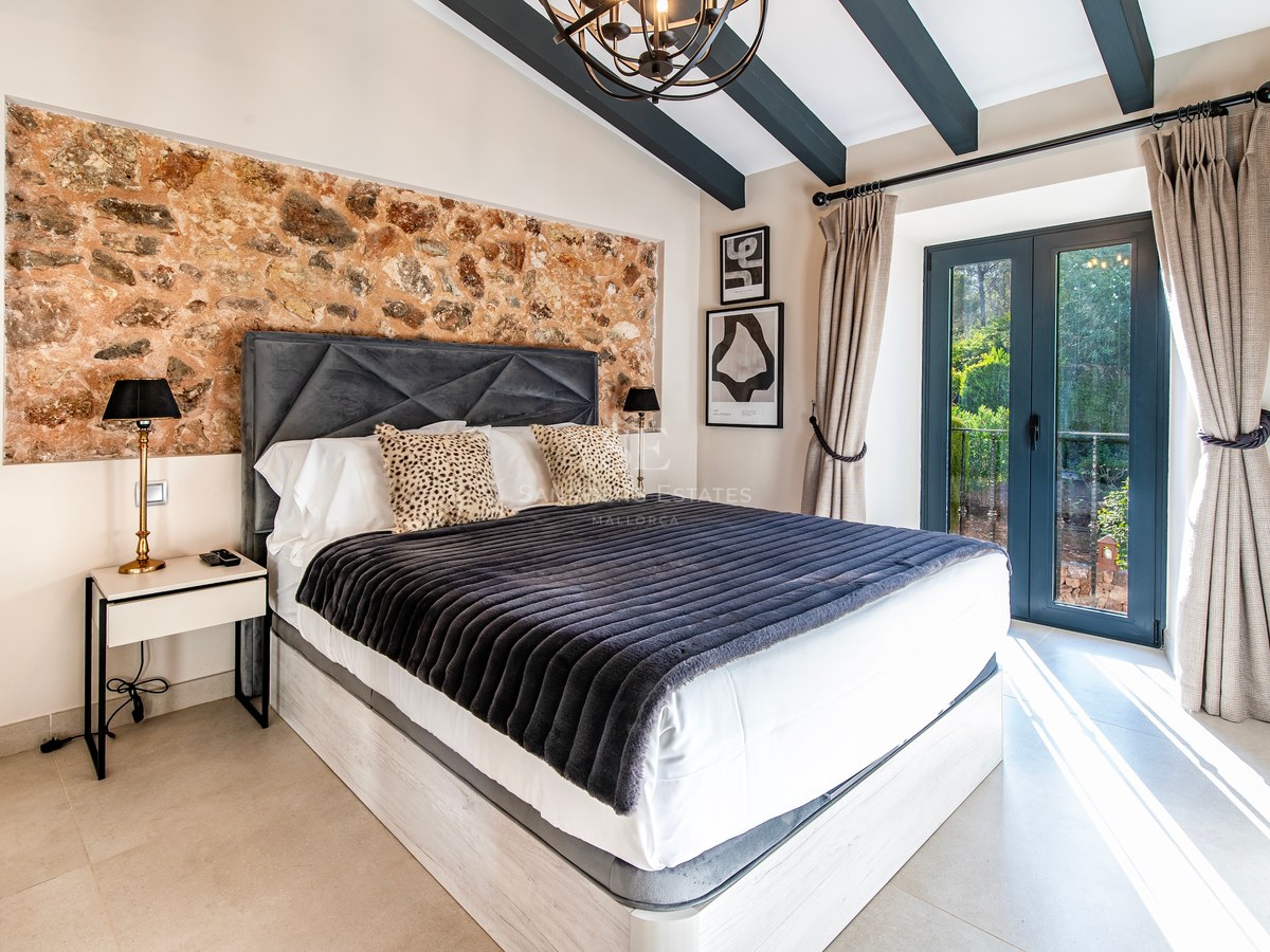Dormitorio de lujo con pared de piedra rústica, vigas oscuras en el techo y puertas francesas al jardín.