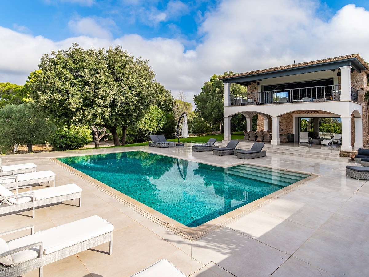 Piscina y terraza de una villa de lujo con fachada de piedra y jardín frondoso bajo un cielo azul.