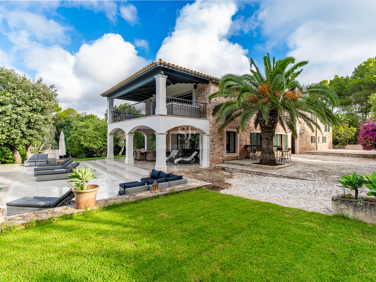 Exterior de una villa de piedra con arcos blancos, una gran palmera, tumbonas y jardín frente a la piscina.
