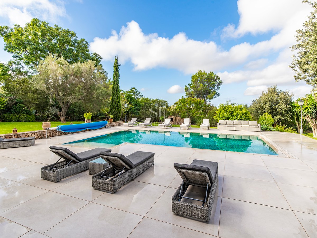 Piscina rectangular azul con amplia terraza de baldosas beige, tumbonas de ratán y jardín frondoso con césped.