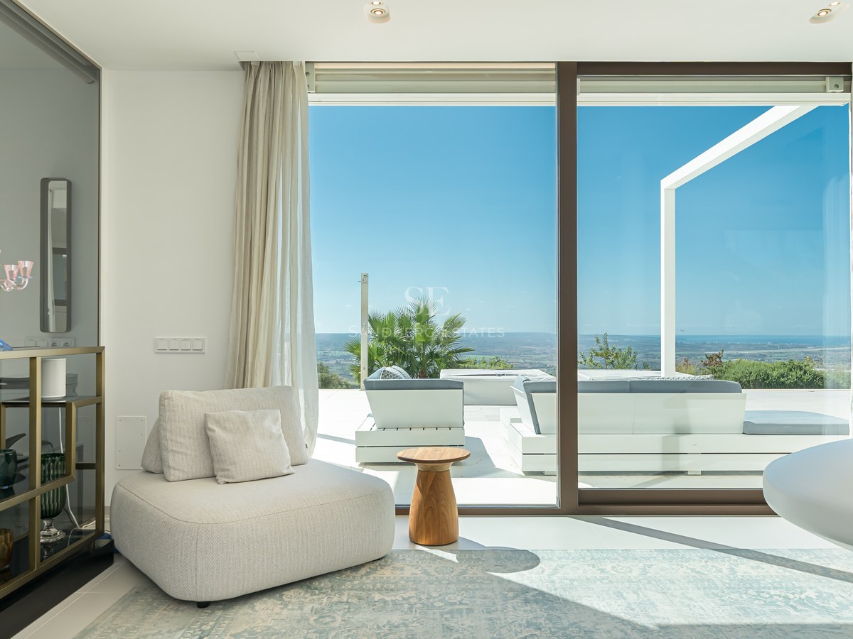 Luminoso salón con ventanales al mar, suelos de madera clara y mobiliario moderno en tonos neutros. Diseño elegante y vistas panorámicas.