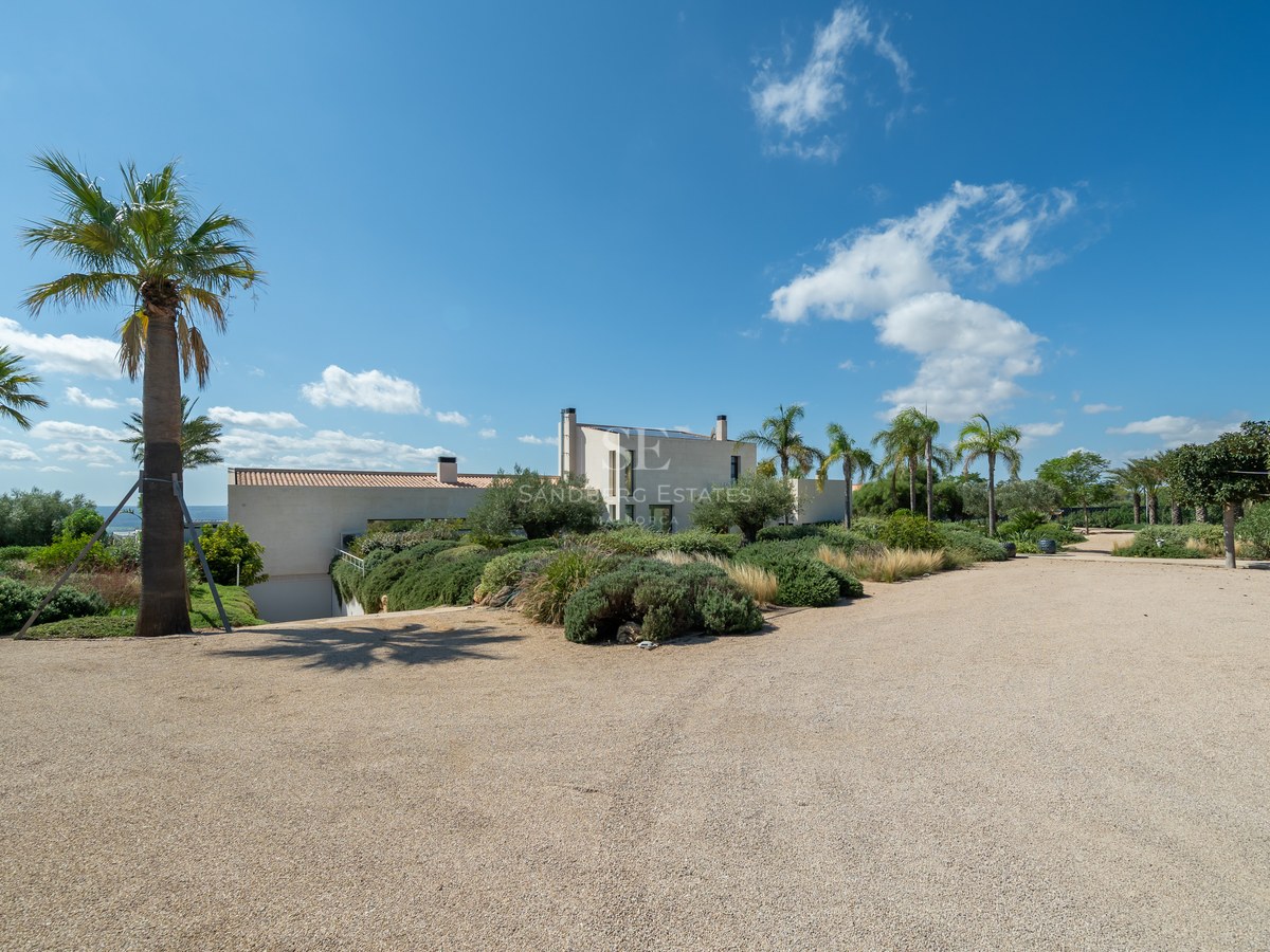 Vista de piscina infinita rodeada de una villa moderna con amplios ventanales y diseño minimalista. Jardín bien cuidado y cielo azul.