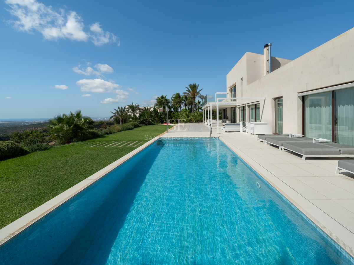 Vista de la piscina con tumbonas, palmeras y una moderna casa de diseño con grandes ventanales. Exterior lujoso y minimalista.