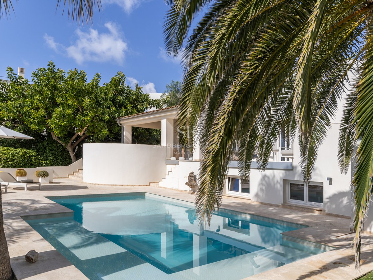 Villa blanche moderne avec piscine rectangulaire entourée de dalles en pierre et de palmiers sous un ciel bleu.