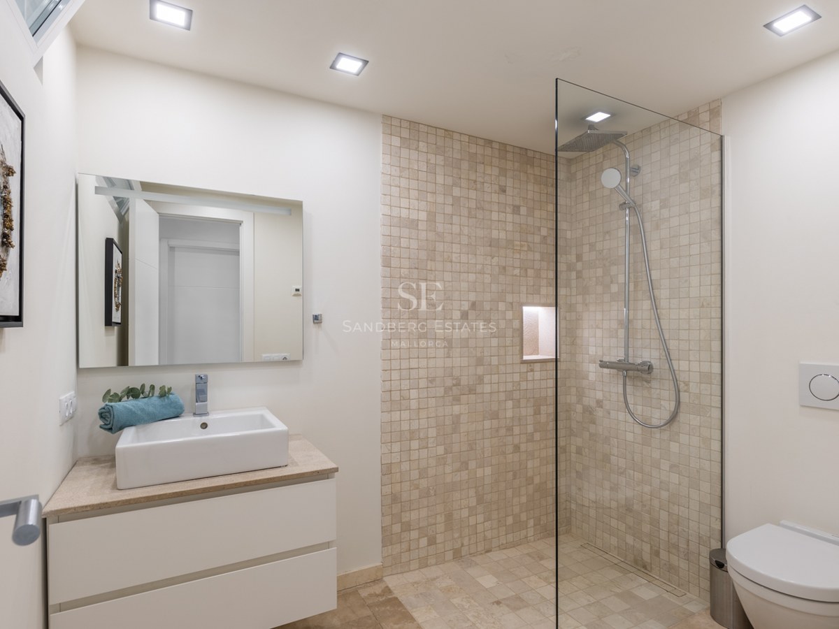 Salle de bains avec douche vitrée, mosaïque en pierre naturelle, meuble vasque blanc et finitions chromées.