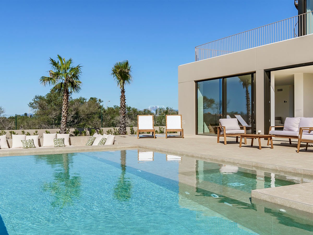 Piscine moderne turquoise avec terrasse en pierre, chaises longues de design et palmiers sous un ciel bleu clair.