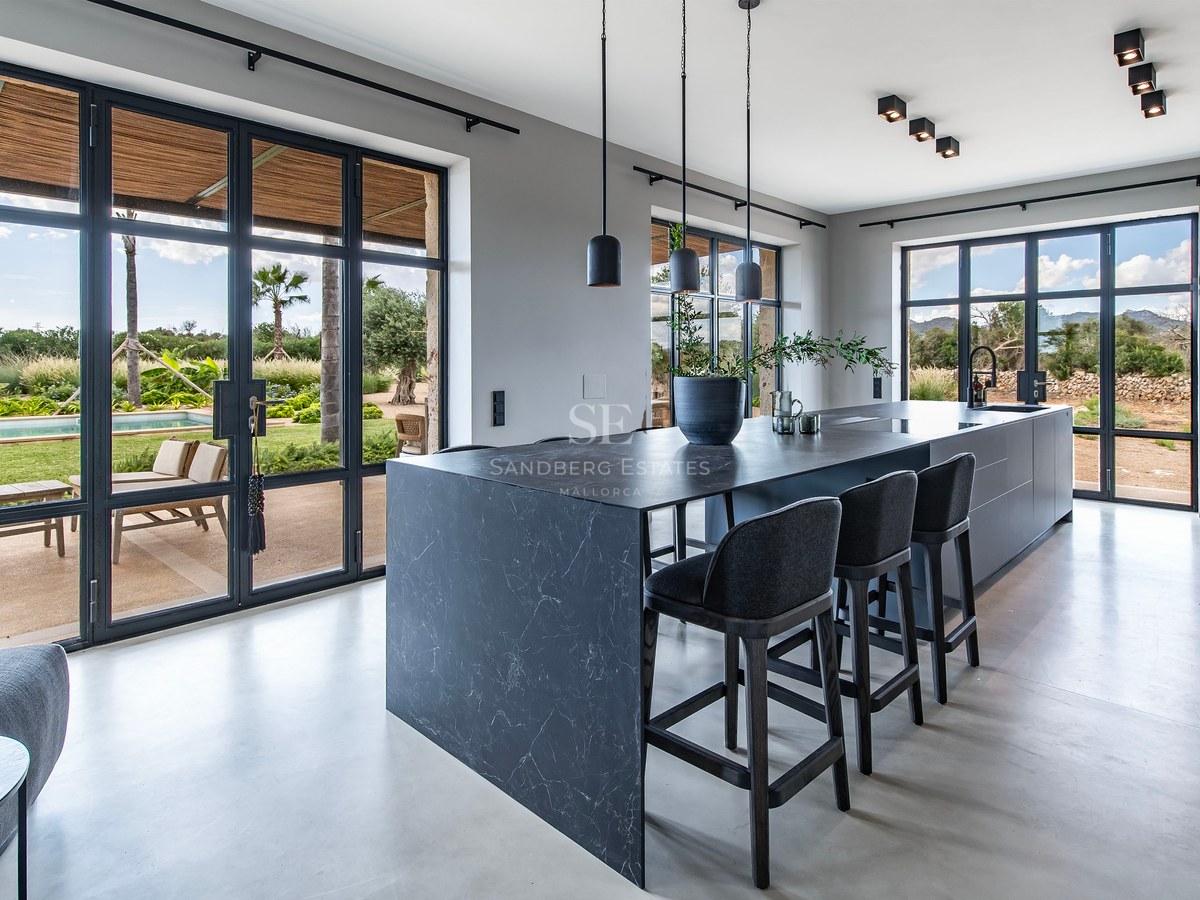 Cuisine moderne avec îlot en marbre noir, tabourets noirs et baies vitrées donnant sur un jardin et une piscine.