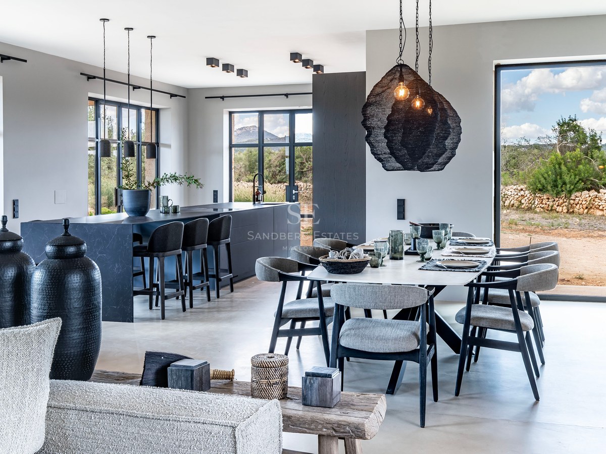 Salle à manger élégante avec une longue table, un îlot de cuisine sombre et de grandes fenêtres donnant sur la campagne.
