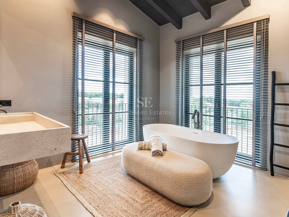 Salle de bain moderne avec baignoire îlot blanche, lavabo en pierre et grandes fenêtres à stores gris.