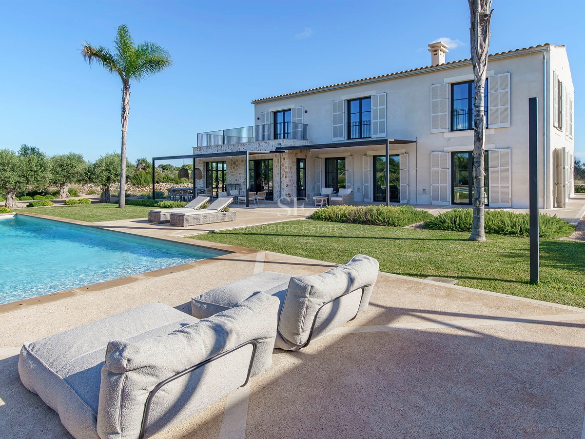 Grande piscine turquoise devant une villa en pierre avec palmiers et pelouse sous un ciel bleu azur.