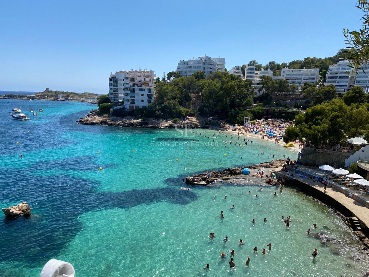 Vista elevada de una bahía turquesa con bañistas, edificios blancos en la costa y acantilados con pinos.