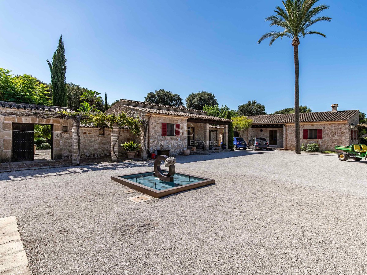 Villa traditionnelle en pierre avec volets rouges, cour en gravier et fontaine moderne sous un ciel bleu.