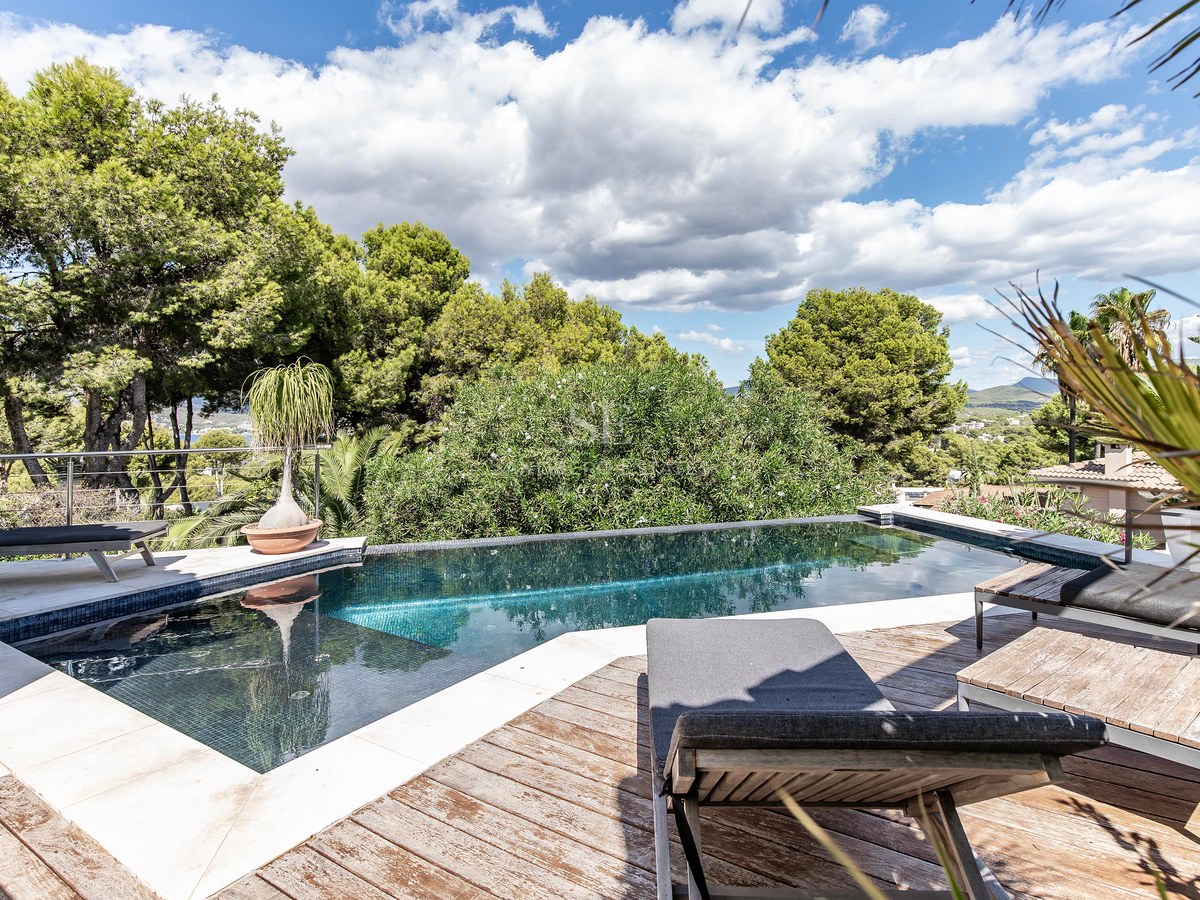 Une piscine à débordement aux carreaux sombres entourée d'une terrasse en bois et de bains de soleil.