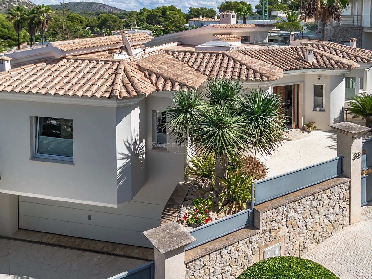 Vue en plongée d'une villa blanche avec toit en tuiles, murs en pierre et jardin tropical sous le soleil.