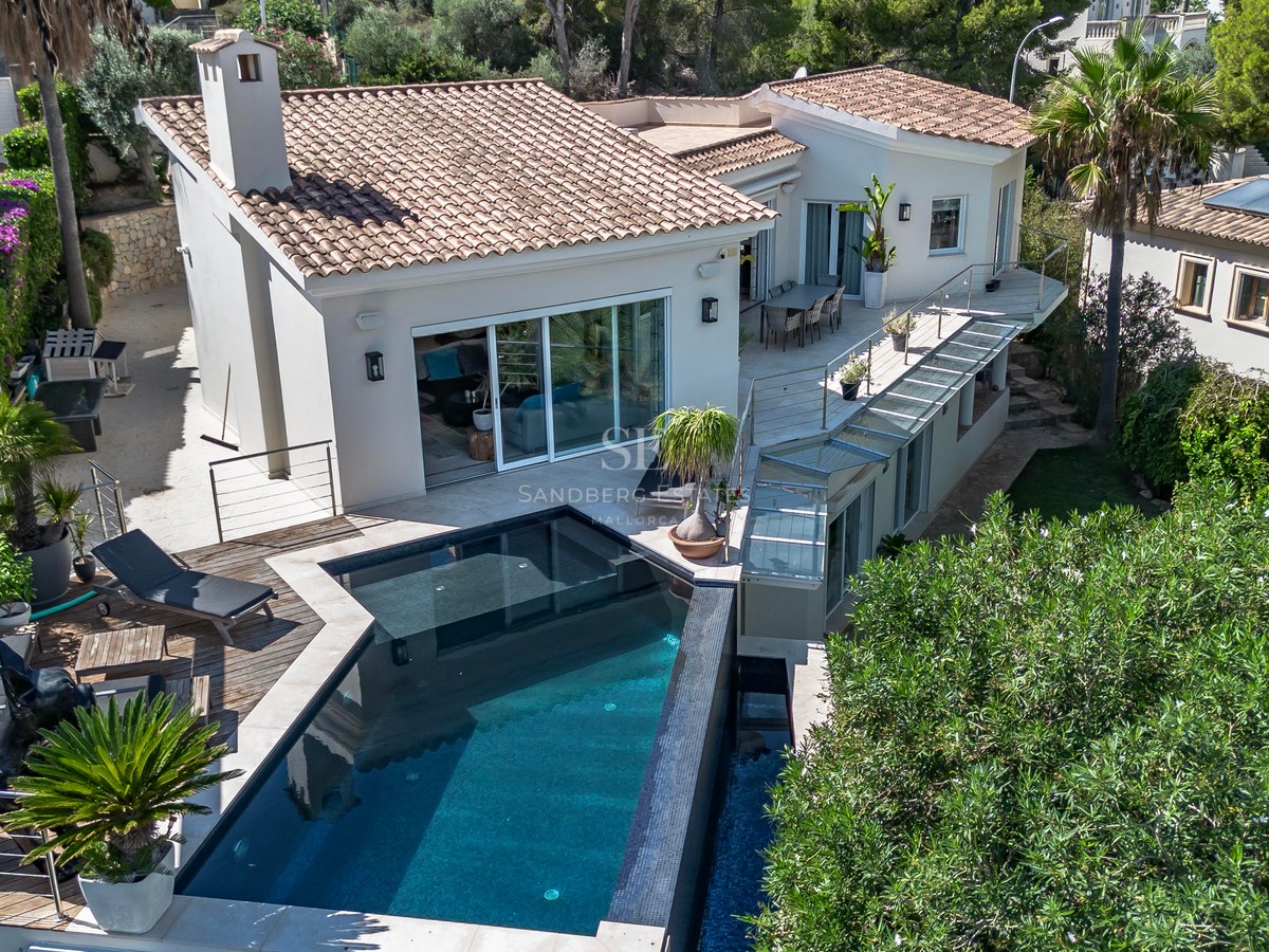 Vue aérienne d'une villa blanche au toit de tuiles avec piscine à débordement et terrasses en bois naturel.