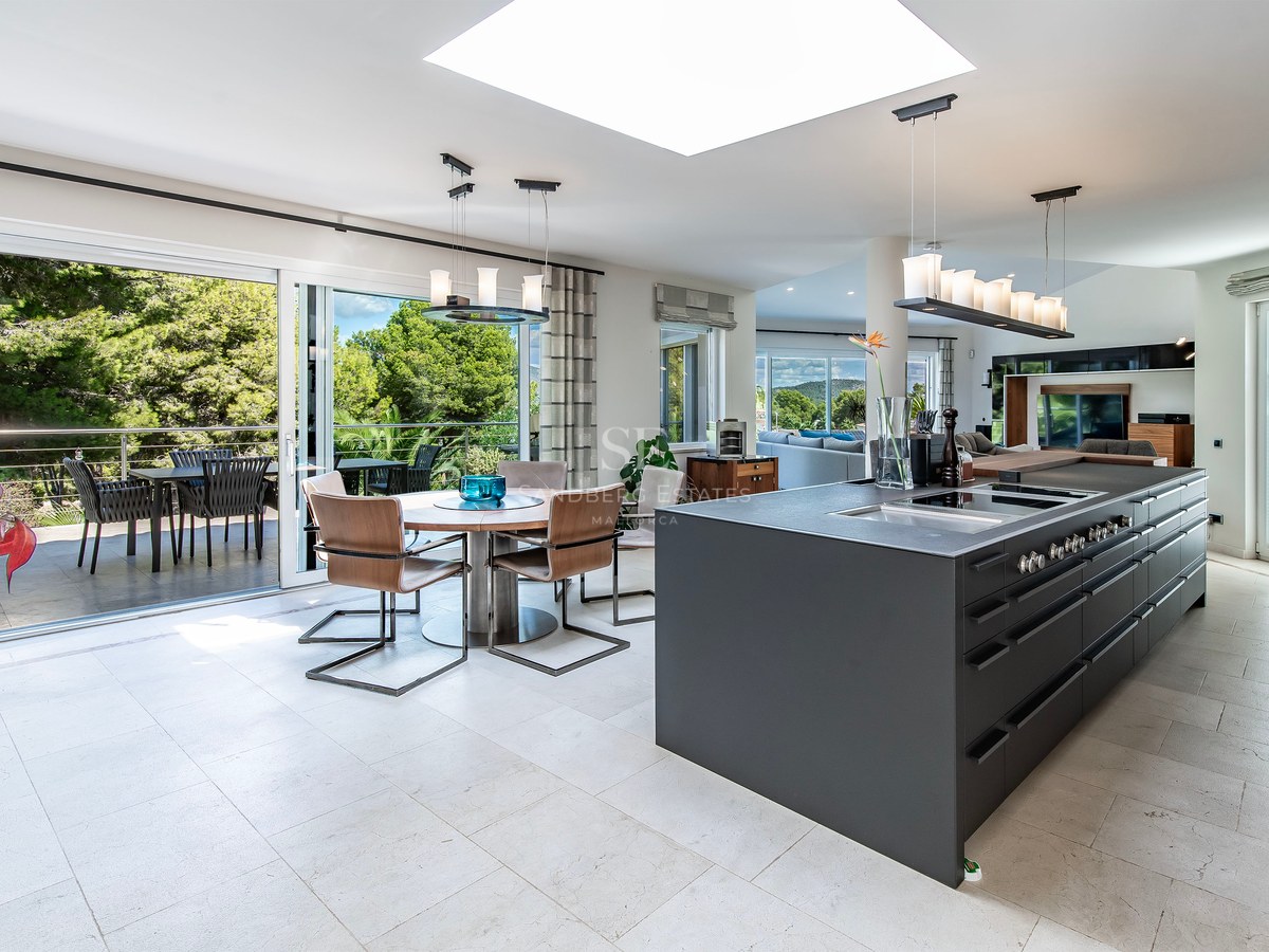 Cuisine moderne avec îlot noir mat, coin repas et grandes baies vitrées ouvrant sur une terrasse boisée.
