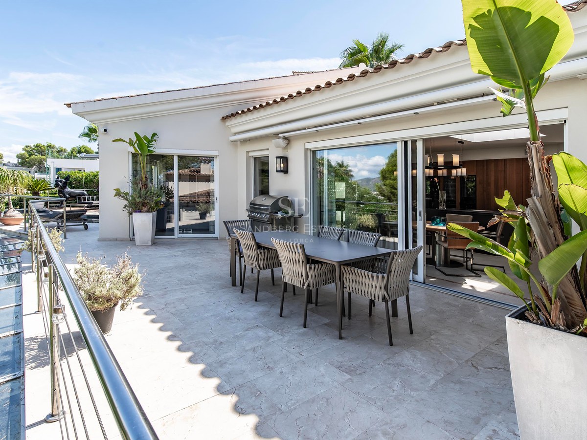 Terrasse ensoleillée avec dallage en pierre, table à manger et barbecue, s'ouvrant sur l'intérieur par des baies vitrées.