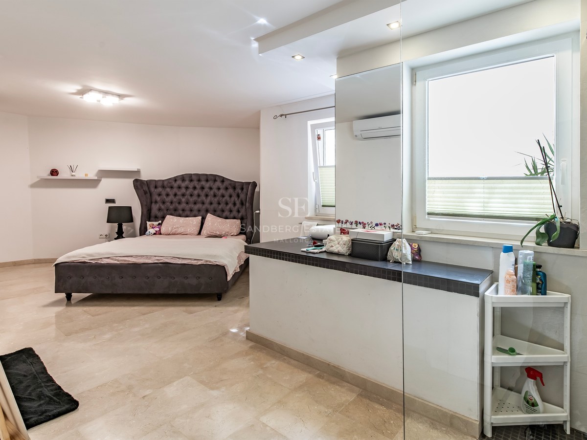 Grande chambre avec lit capitonné gris anthracite, sol en pierre crème et cloison avec rangement.