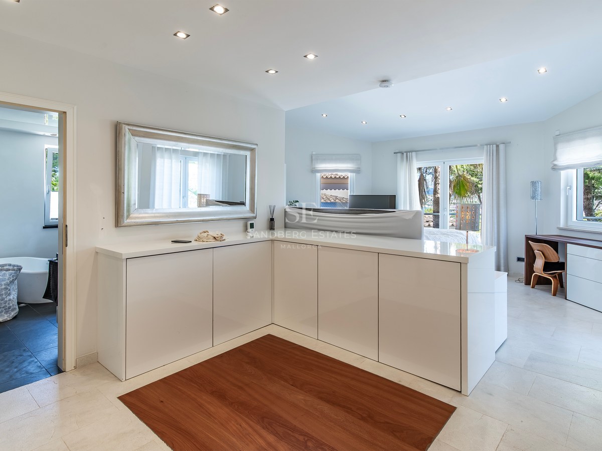 Grande chambre moderne avec rangements blancs, grand miroir, sol en pierre et vue sur la salle de bains attenante.