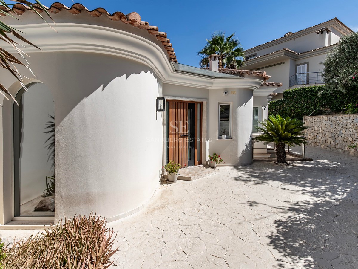 Extérieur de villa blanche aux lignes courbes, porte en bois, toit en terre cuite et allée pavée sous ciel bleu.