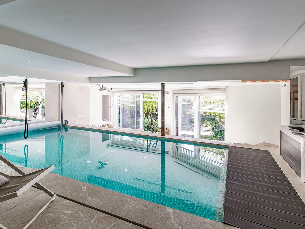 Piscina interior con mosaicos turquesa, jacuzzi y puertas de cristal hacia el jardín.