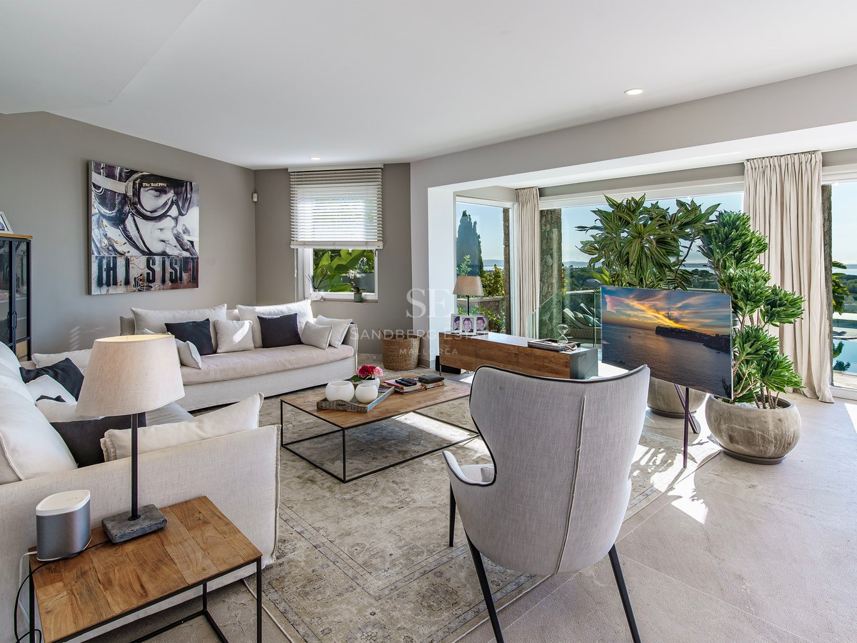 Salón moderno con sofás blancos, grandes ventanales con vistas a la piscina y al mar Mediterráneo.