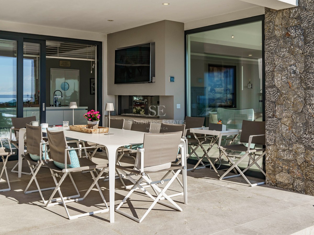 Terraza cubierta moderna con mesa blanca, sillas grises, pared de piedra natural y puertas de cristal con vistas al mar.