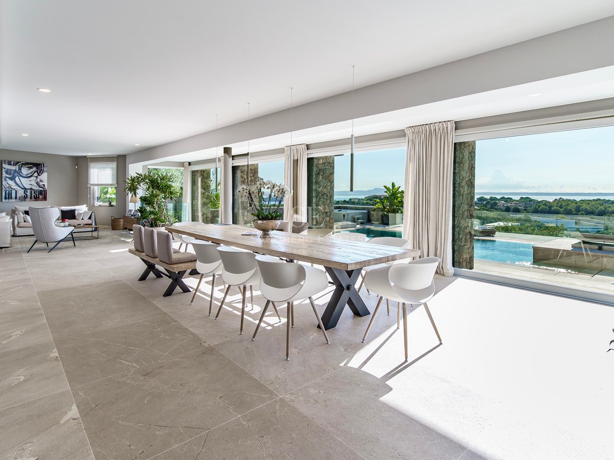 Comedor de planta abierta con mesa de madera y ventanales de suelo a techo con vistas a la piscina y al mar.