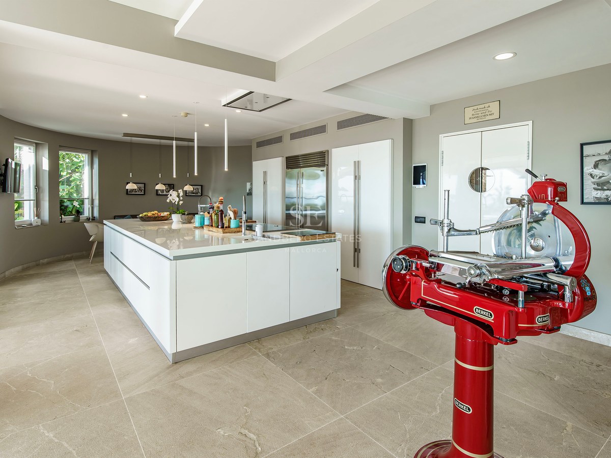Cocina moderna blanca con isla grande, suelo de piedra, electrodomésticos integrados y cortadora de fiambre roja.