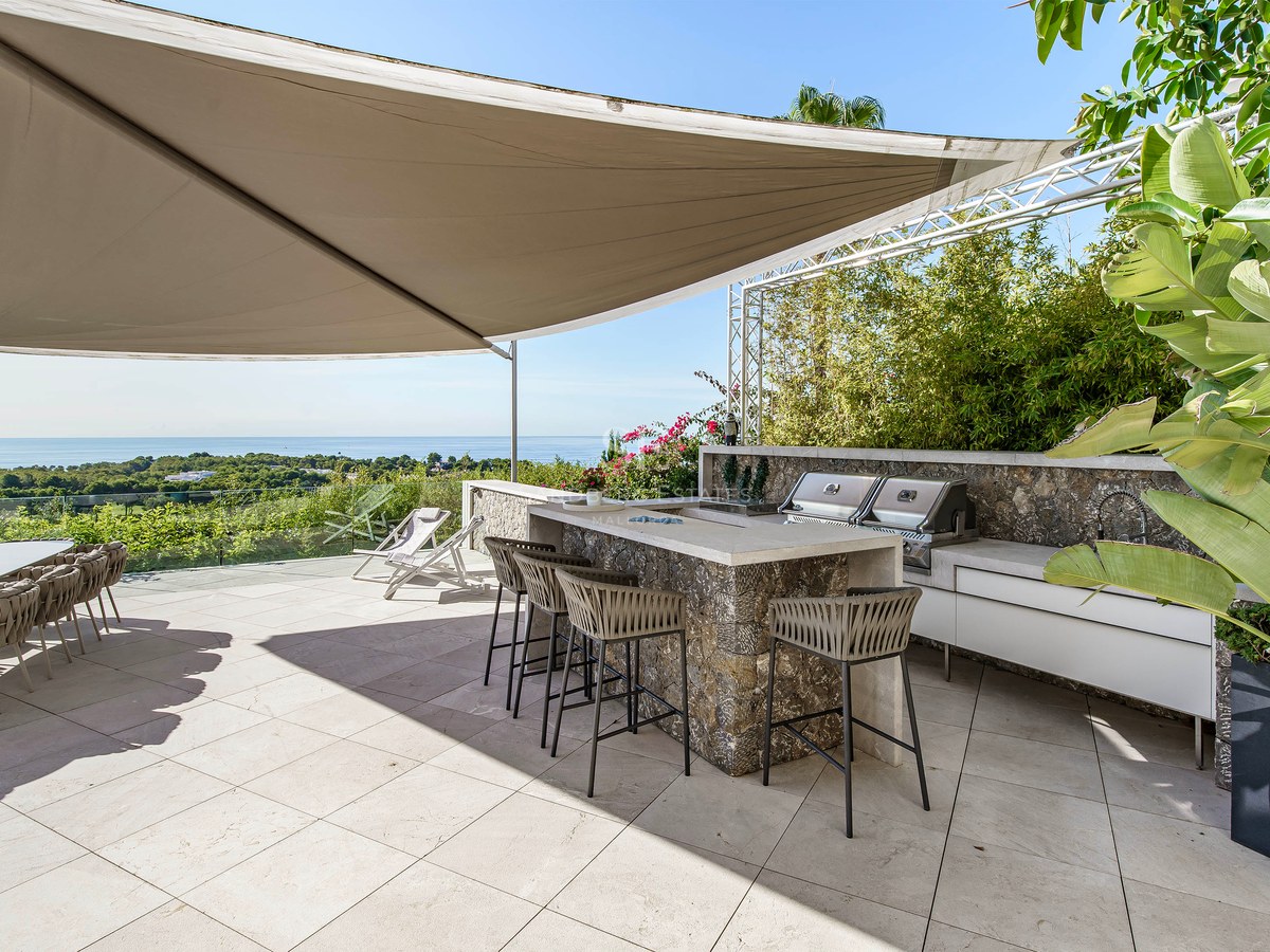 Cocina exterior moderna en terraza de piedra con barbacoa, barra y vela de sombra con vistas al mar.