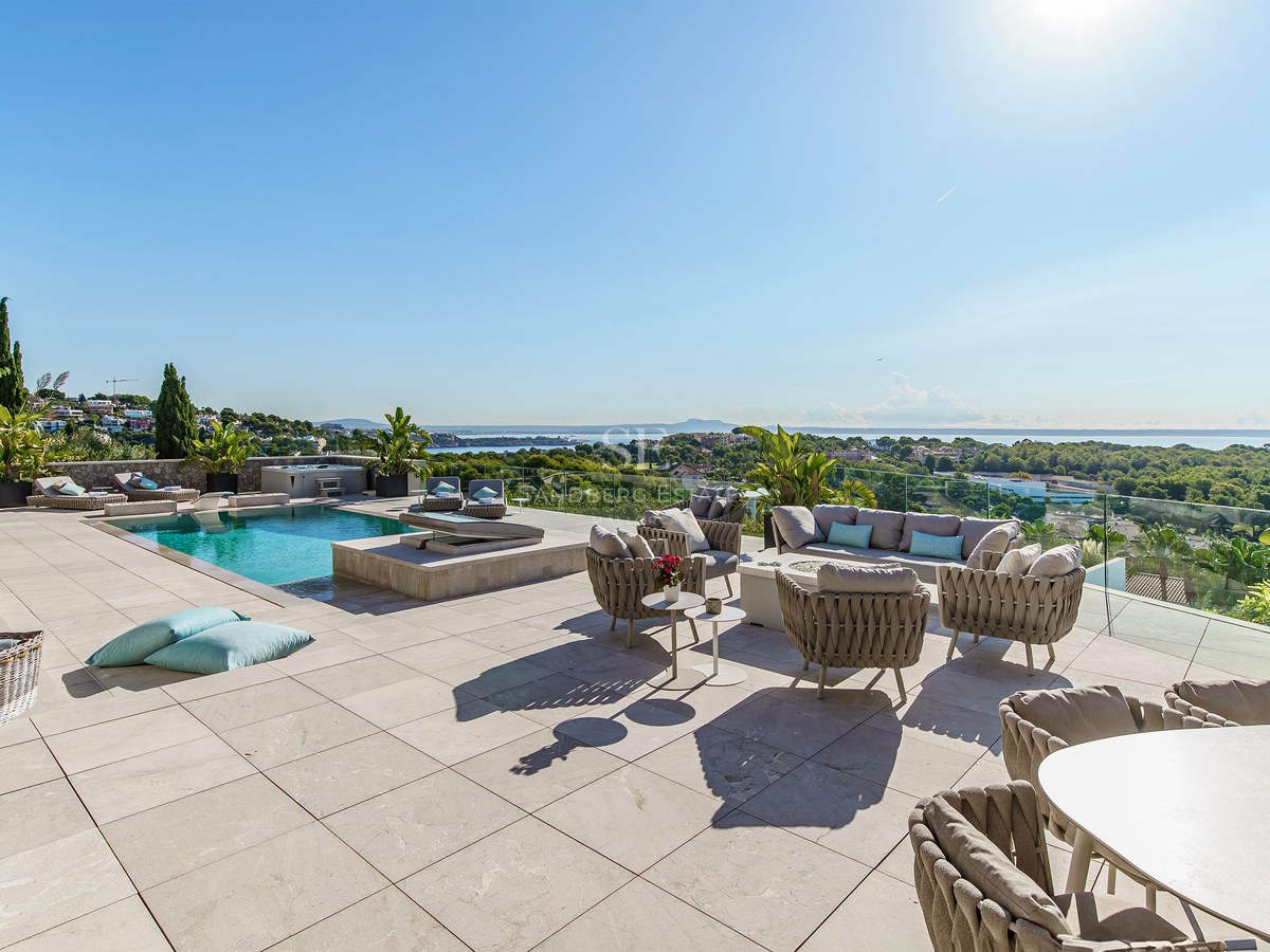 Terraza de piedra con muebles de diseño, piscina y vistas panorámicas al mar y al paisaje verde bajo un cielo despejado.