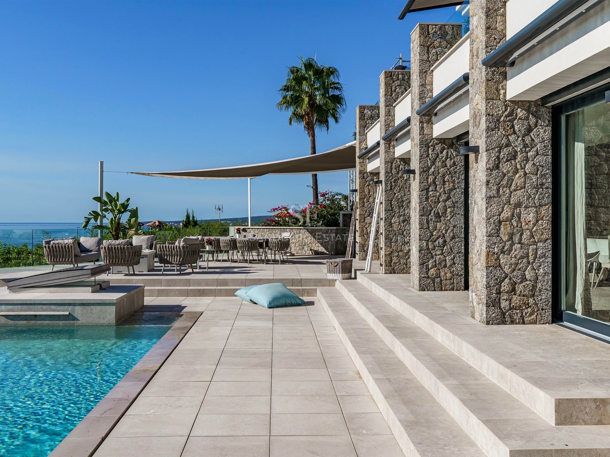 Terraza de piedra con piscina infinity, zona de estar sombreada y vistas panorámicas al mar bajo un cielo despejado.