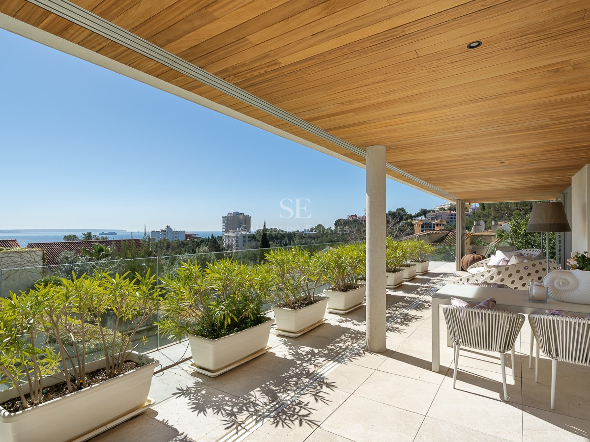 A spacious covered terrace with a wooden slatted ceiling, white planters, and a dining area overlooking the sea.