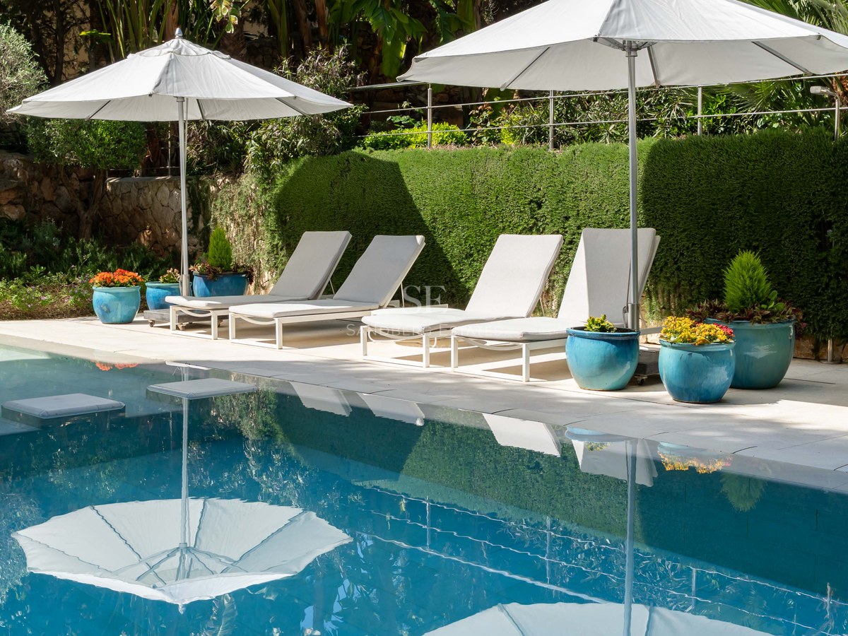 Sun loungers with white umbrellas beside a blue swimming pool, framed by lush green hedges and turquoise planters.