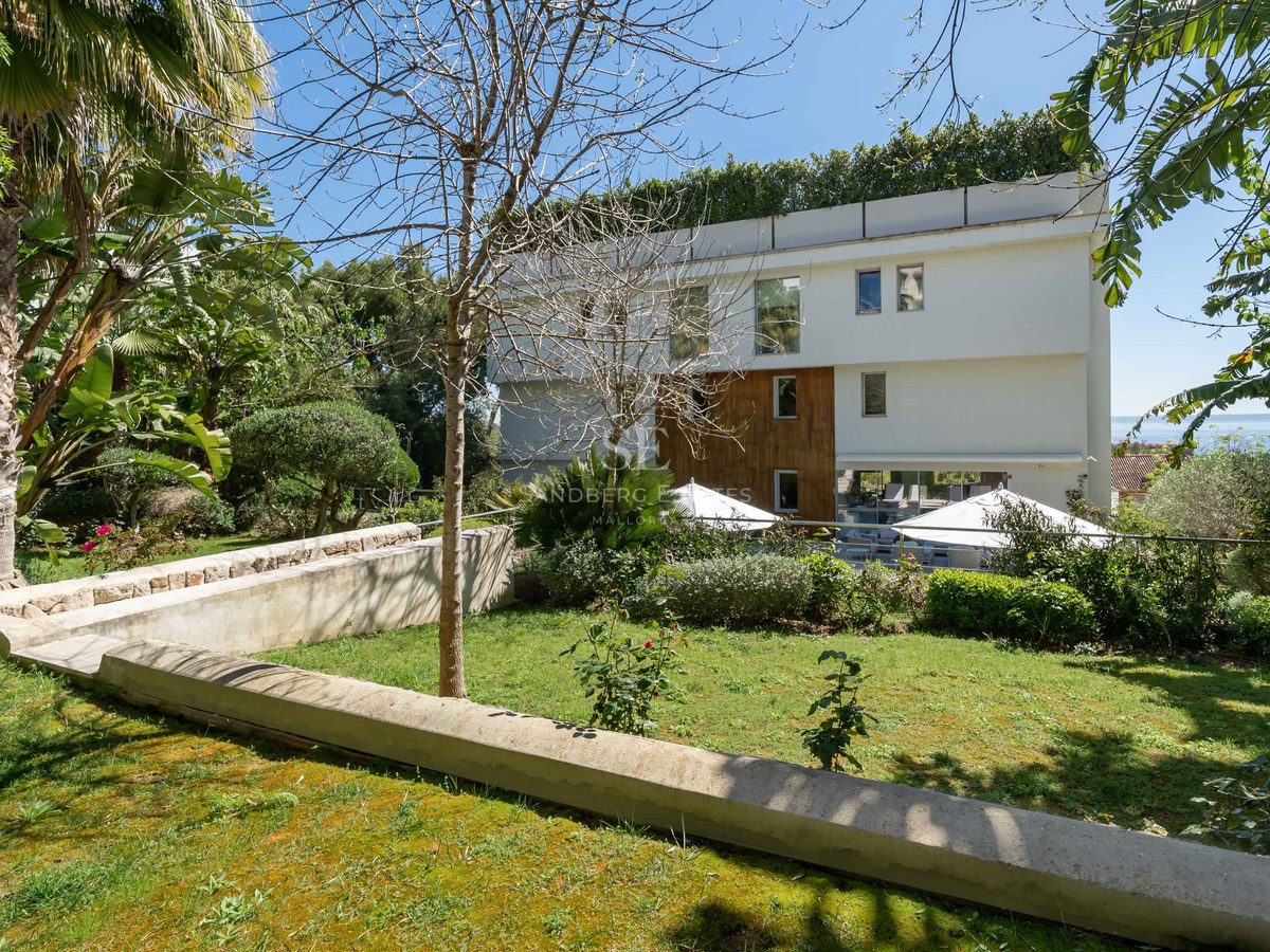 Modern white three-story villa with wood paneling accents viewed from a manicured green garden with stone walls.
