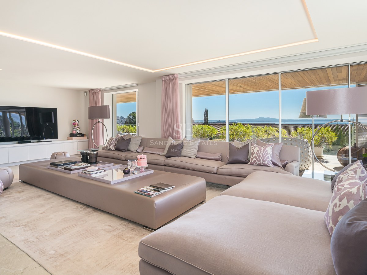 Spacious modern living room with large modular taupe sofas, natural stone flooring, and floor-to-ceiling windows with sea views.