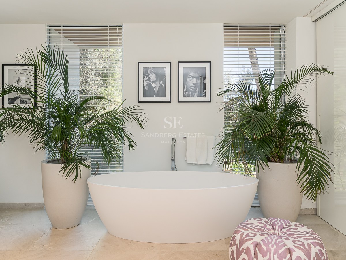 Modern master bathroom featuring a white freestanding oval bathtub flanked by large potted palms and natural light.