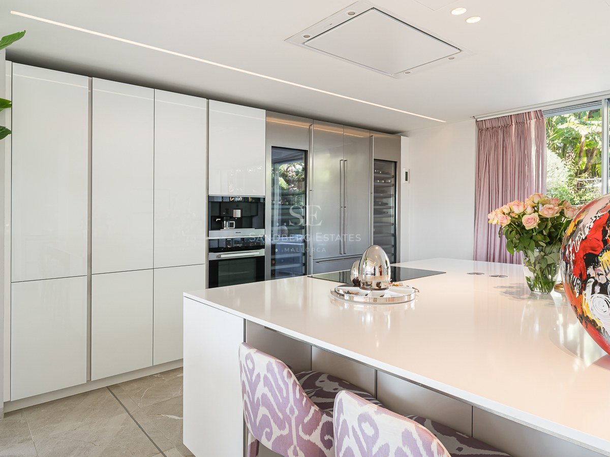 Bright modern kitchen featuring a white central island, floor-to-ceiling cabinetry, and large windows with garden views.