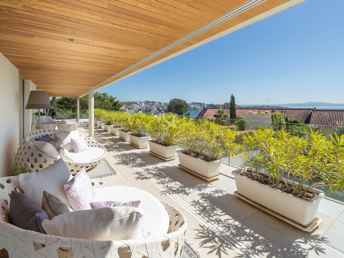 Modern covered terrace with wooden ceiling, white lounge furniture, and potted plants overlooking the sea.
