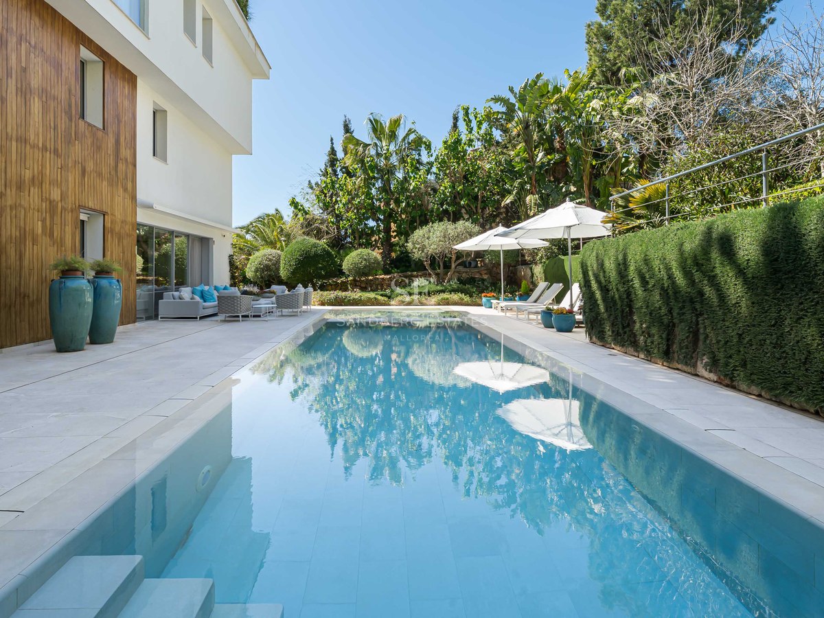 A long turquoise swimming pool surrounded by a cream stone terrace, wood-paneled walls, and vibrant green hedges.