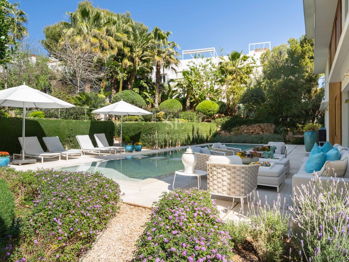 A sunny private swimming pool area with white lounge furniture, umbrellas, and mature green landscaping.