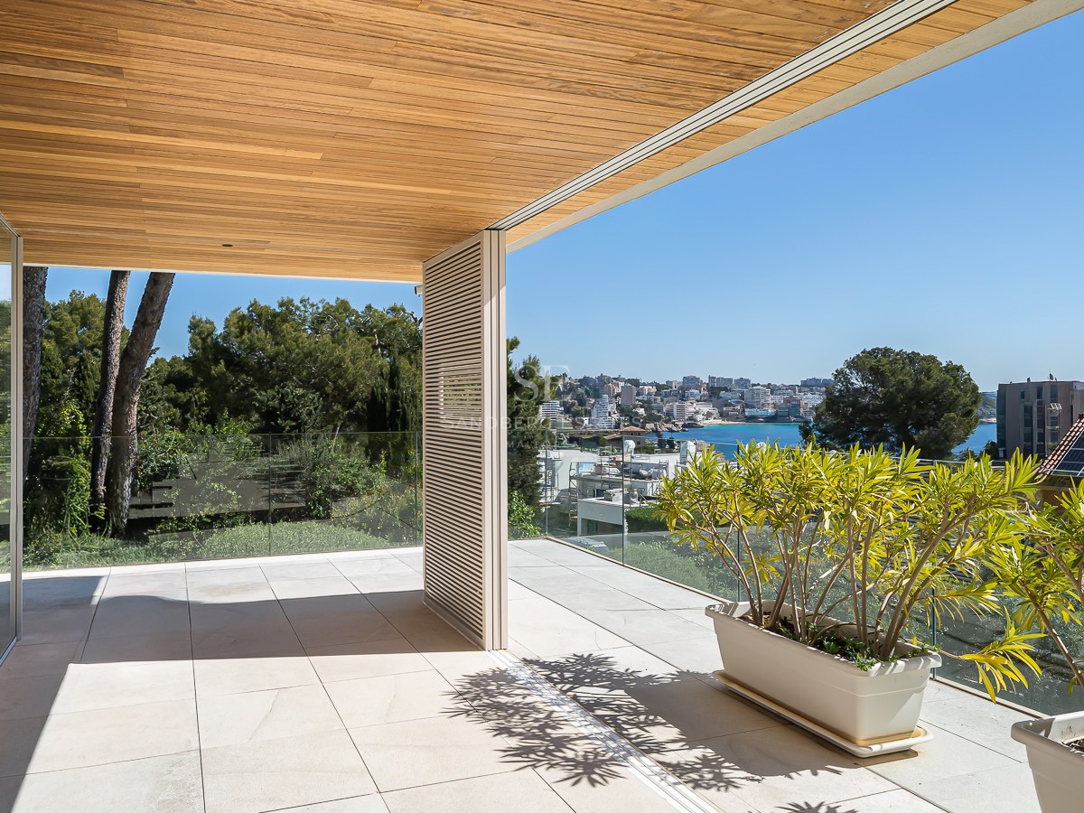 Sun-drenched modern terrace with natural stone flooring, glass railings, and a wooden ceiling overlooking a coastal town.