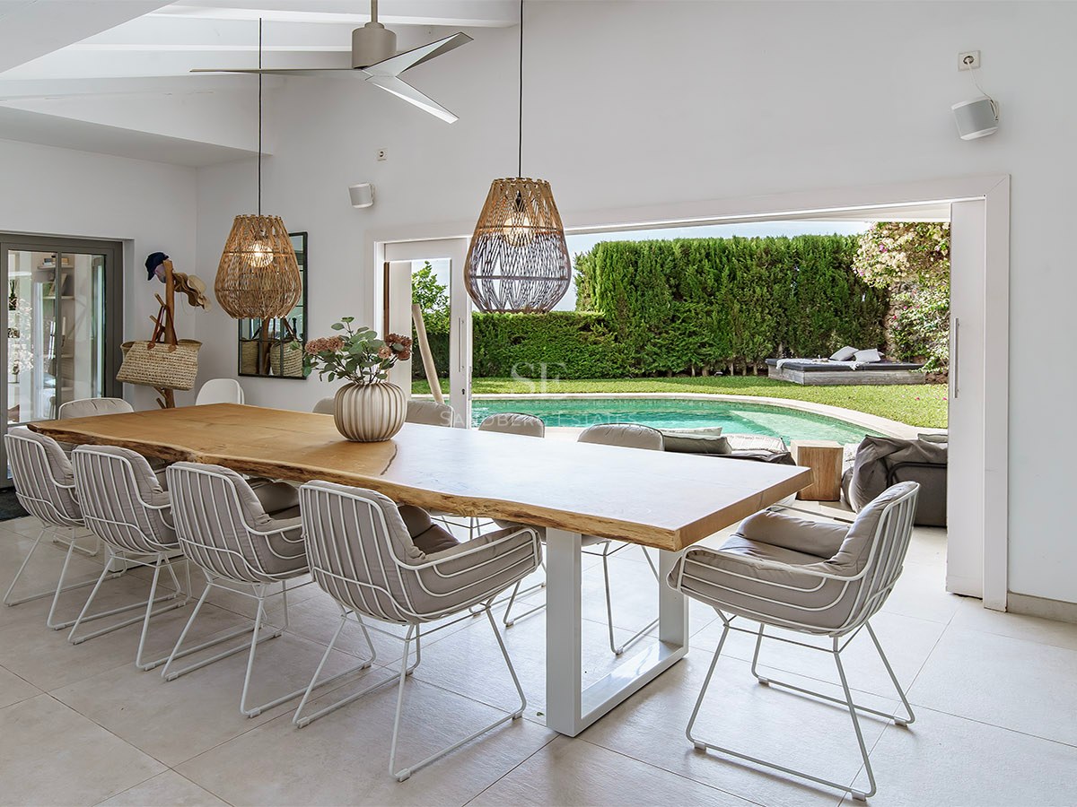 Elegante comedor con gran mesa de madera y lámparas de mimbre, abierto a una terraza con piscina y jardín.
