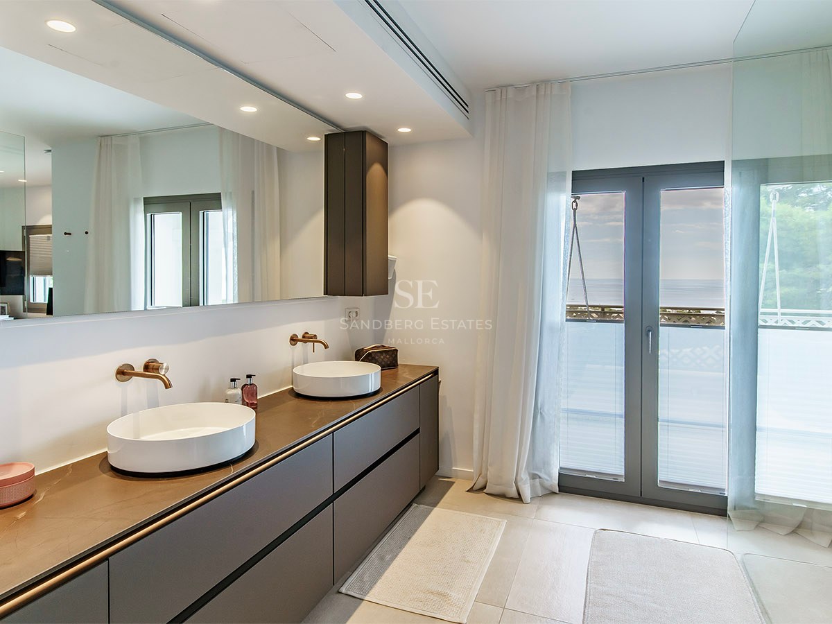 Baño principal moderno con lavabo doble, gran espejo y acceso al balcón con vistas al mar.