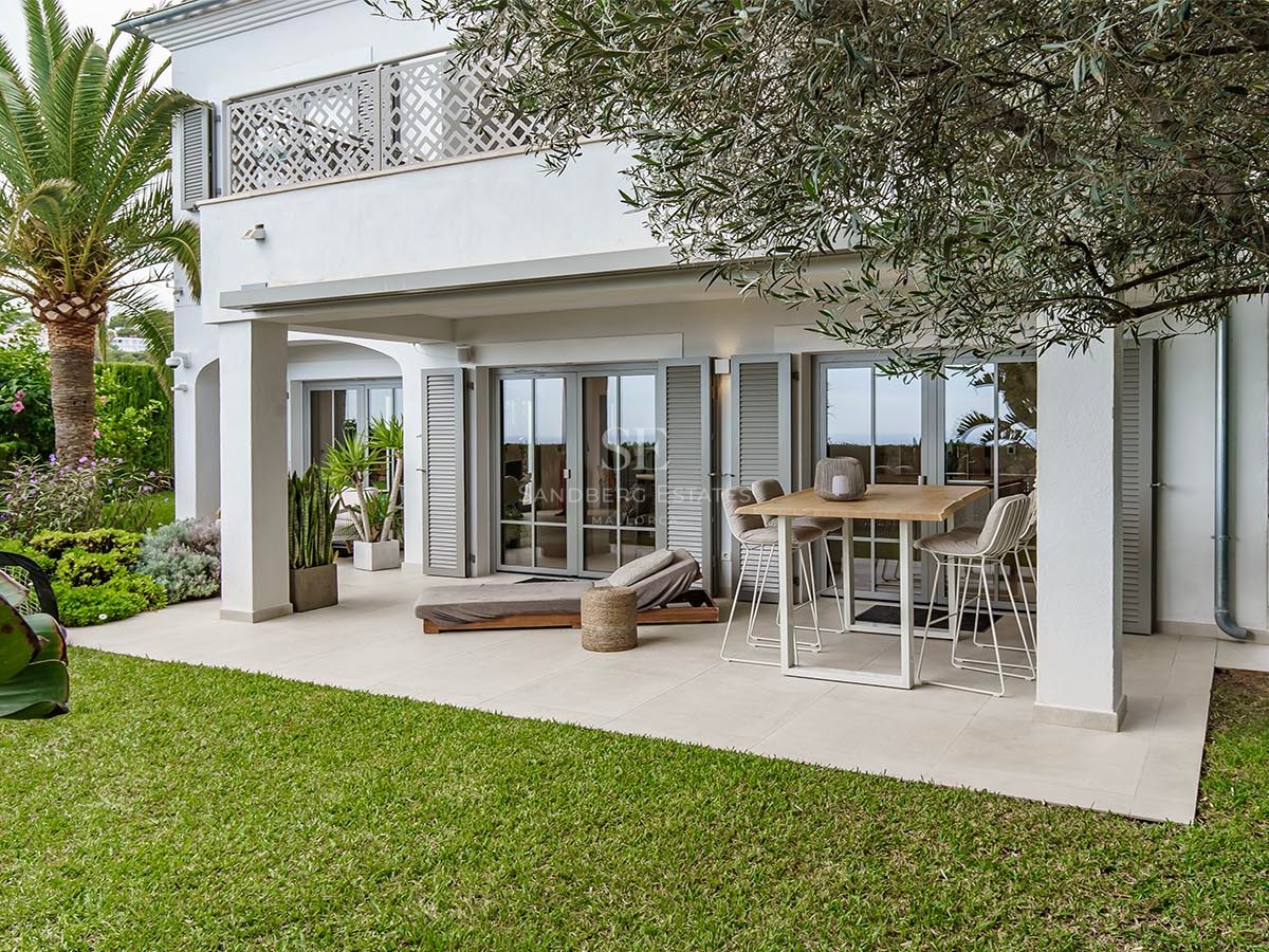 Villa mediterránea blanca con porche cubierto, mesa alta, tumbona, césped verde y palmeras.